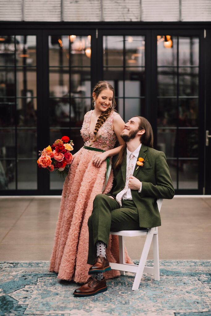 Bride in a blush pink textured gown holding a bold red and orange bouquet while standing beside the groom seated on a white chair inside a modern Madison wedding venue