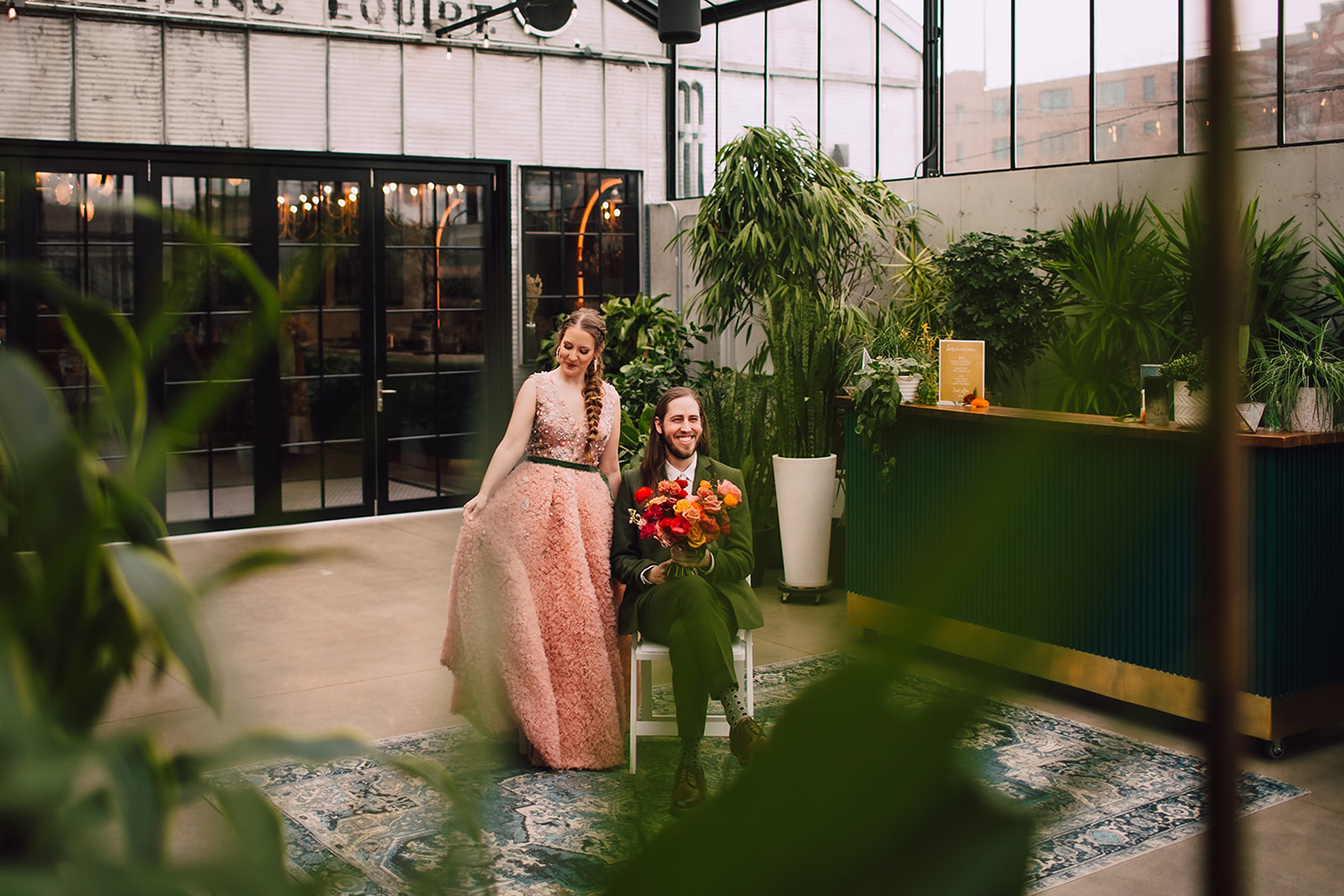 Wedding couple posing together in a light-filled, plant-filled Madison wedding venue with industrial windows, concrete floors, and a modern bar backdrop