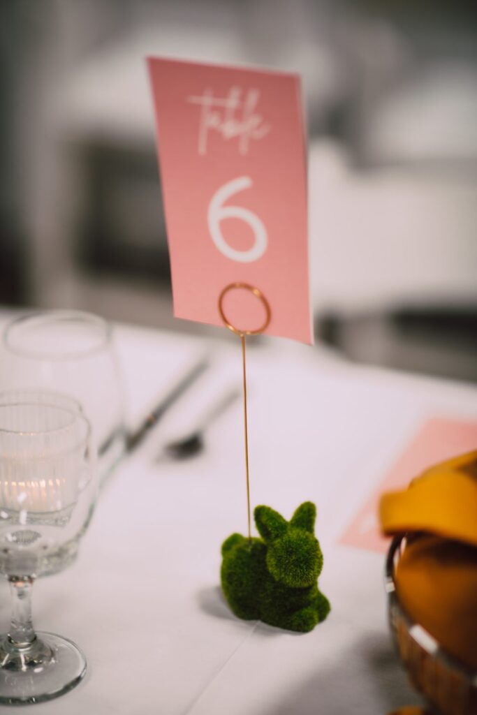 Pink wedding table number card displayed in a gold holder with a moss bunny accent on a white reception table at a Madison wedding venue
