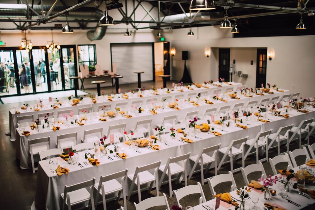 Long banquet-style reception tables with white linens, mustard napkins, and floral accents inside an industrial wedding reception venue in Madison WI