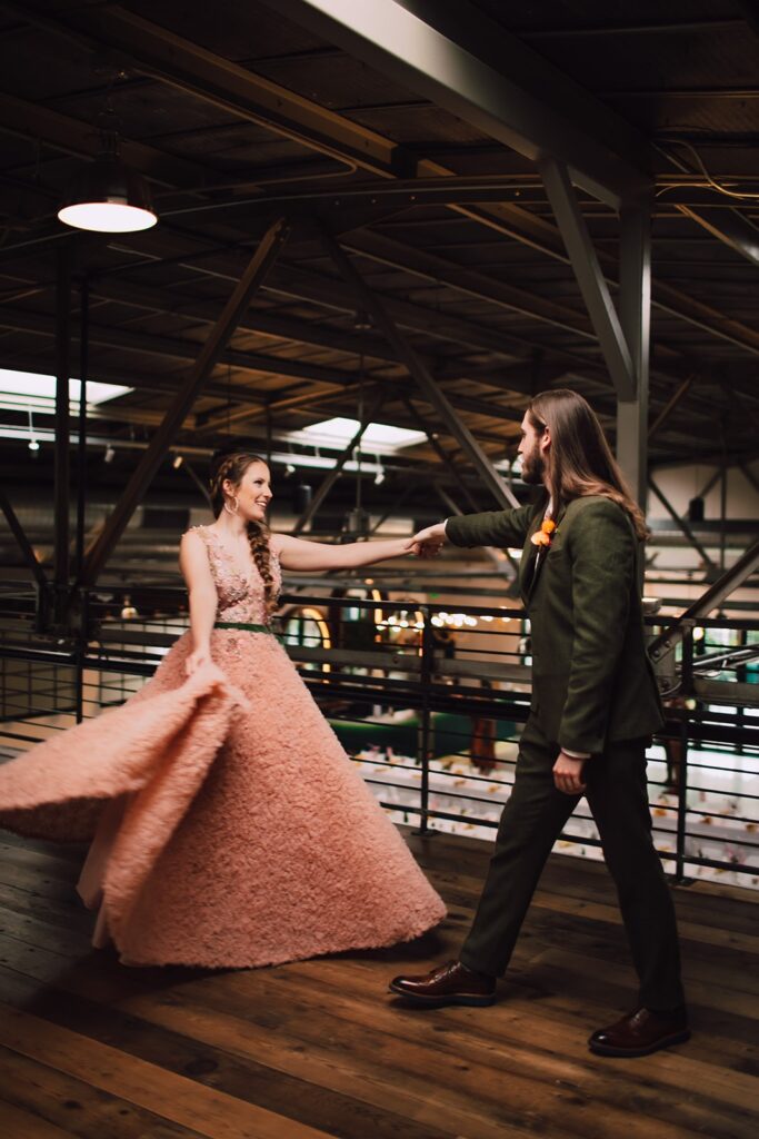 Bride in a blush pink textured gown twirling as the groom reaches for her hand on an industrial mezzanine inside a modern Madison wedding venue, with exposed beams and reception tables below