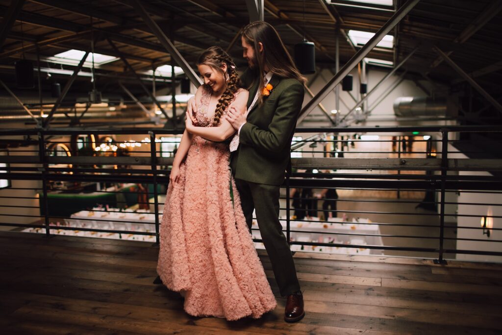 Bride and groom sharing a quiet embrace on an upper-level balcony inside an industrial wedding venue in Madison WI, overlooking the reception space below