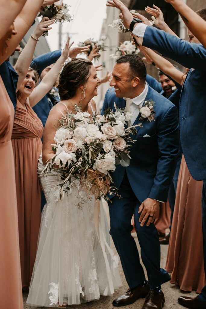 Bride and groom smiling as they walk through a celebratory tunnel of wedding guests holding bouquets during a joyful outdoor wedding at one of the most memorable wedding venues in Madison
