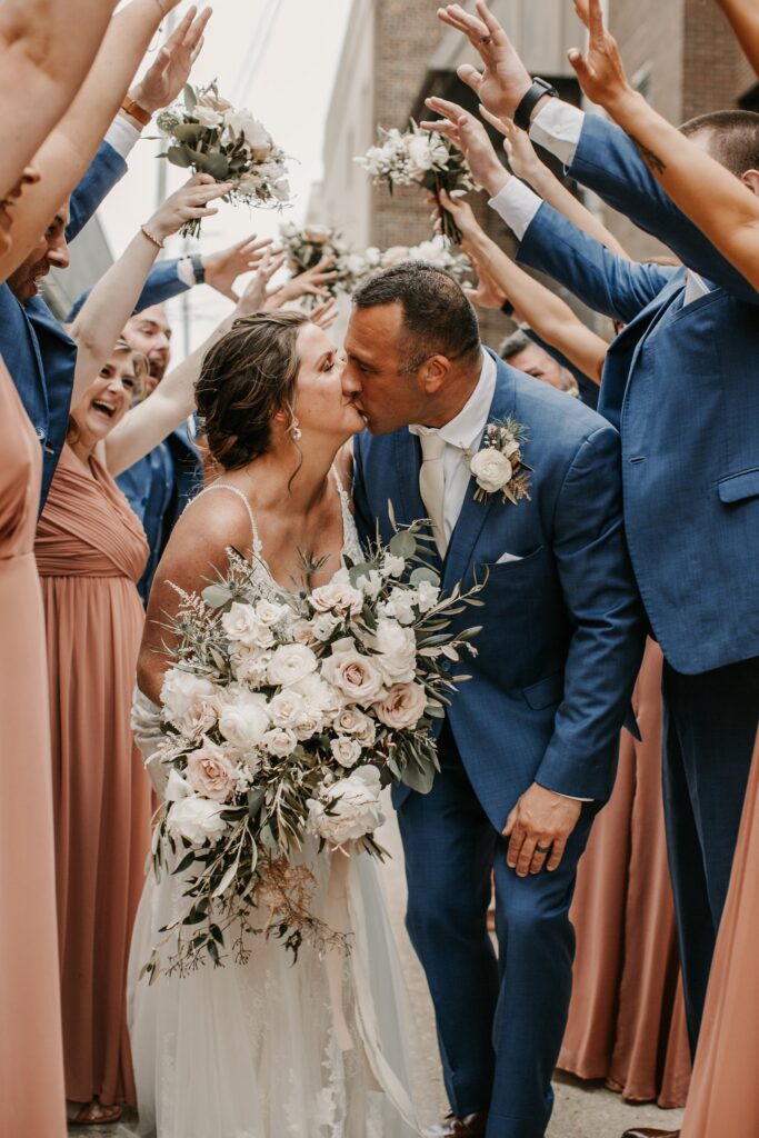 Bride and groom sharing a kiss while guests cheer and raise bouquets around them during a lively outdoor celebration at a Madison wedding venue