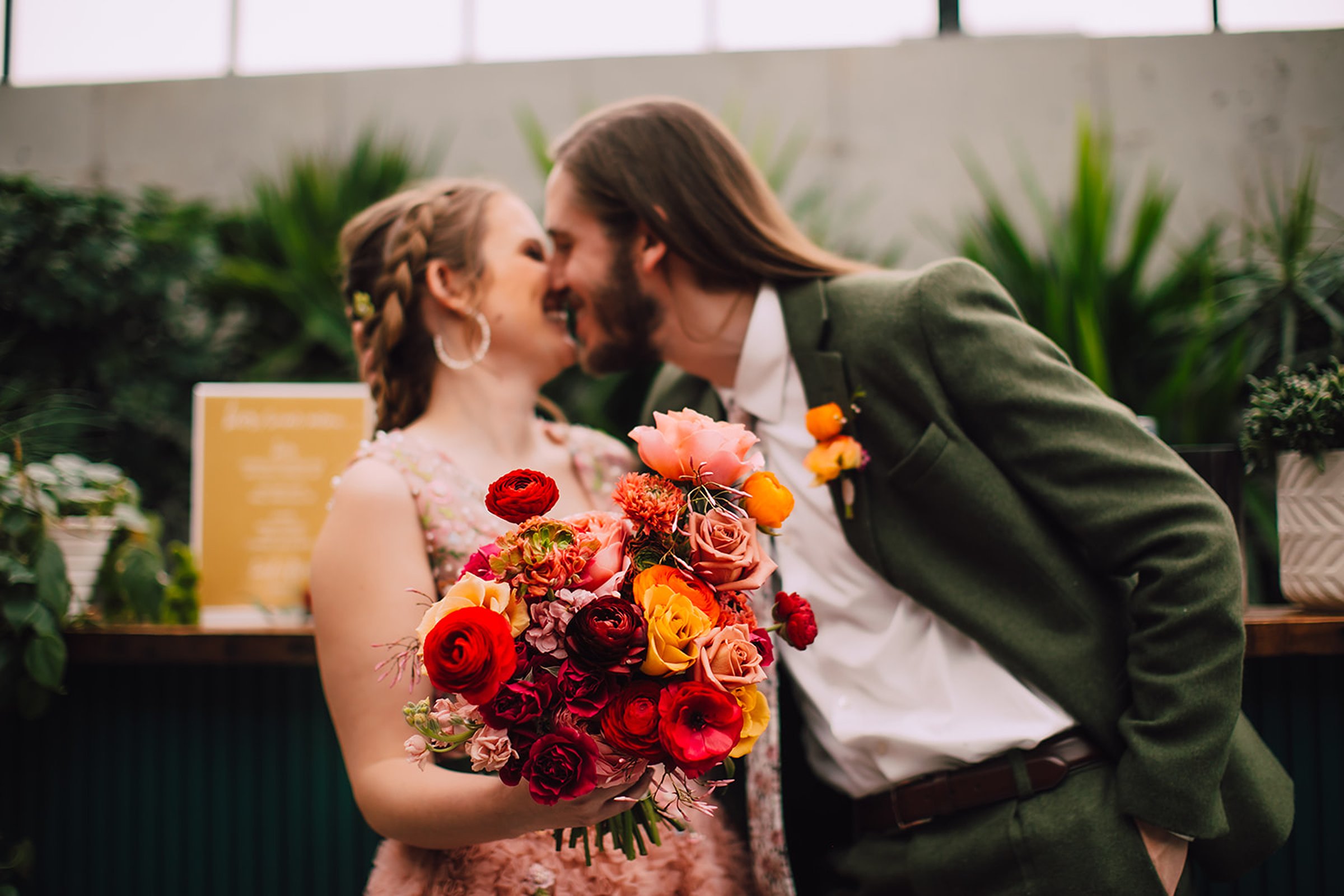 Bride holding a vibrant bouquet of red, orange, and pink flowers while sharing a kiss with the groom inside a modern Madison wedding venue surrounded by greenery