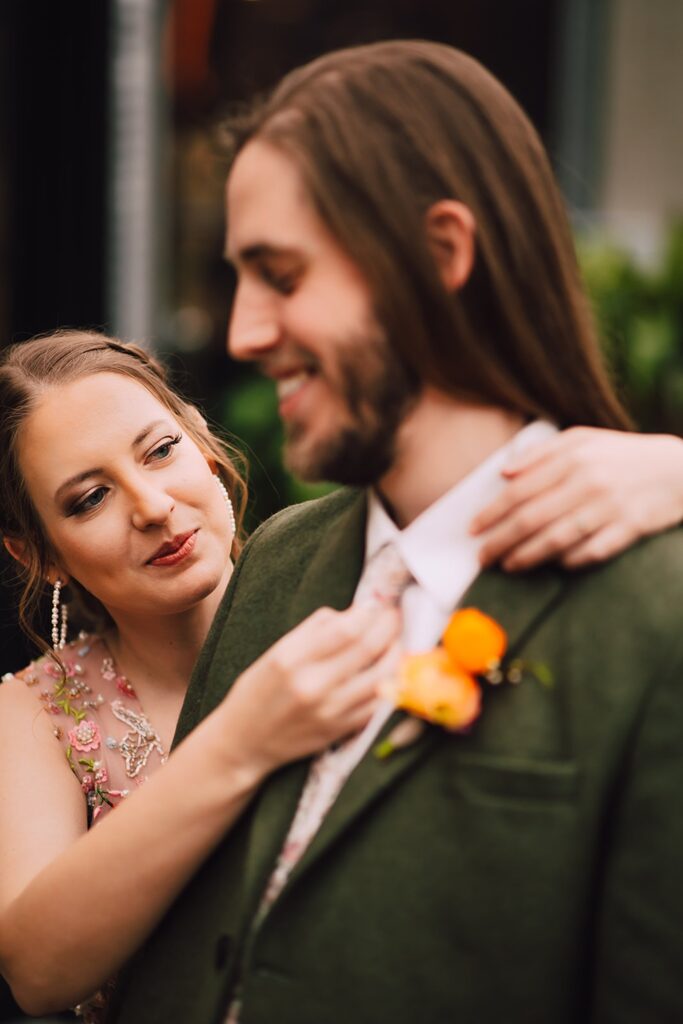Bride adjusting the groom’s floral tie while standing together inside a lush, plant-filled wedding venue in Madison WI