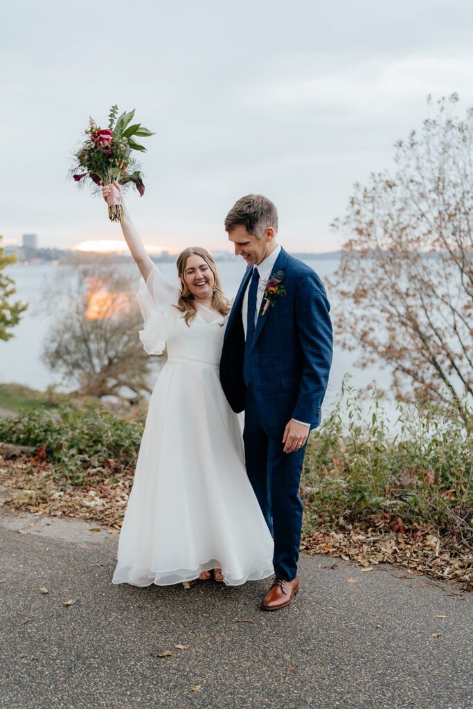 Bride raising her bouquet while standing with the groom by a lakeside path, capturing a joyful moment from getting married in Madison WI and scenic places to get married in Madison WI.