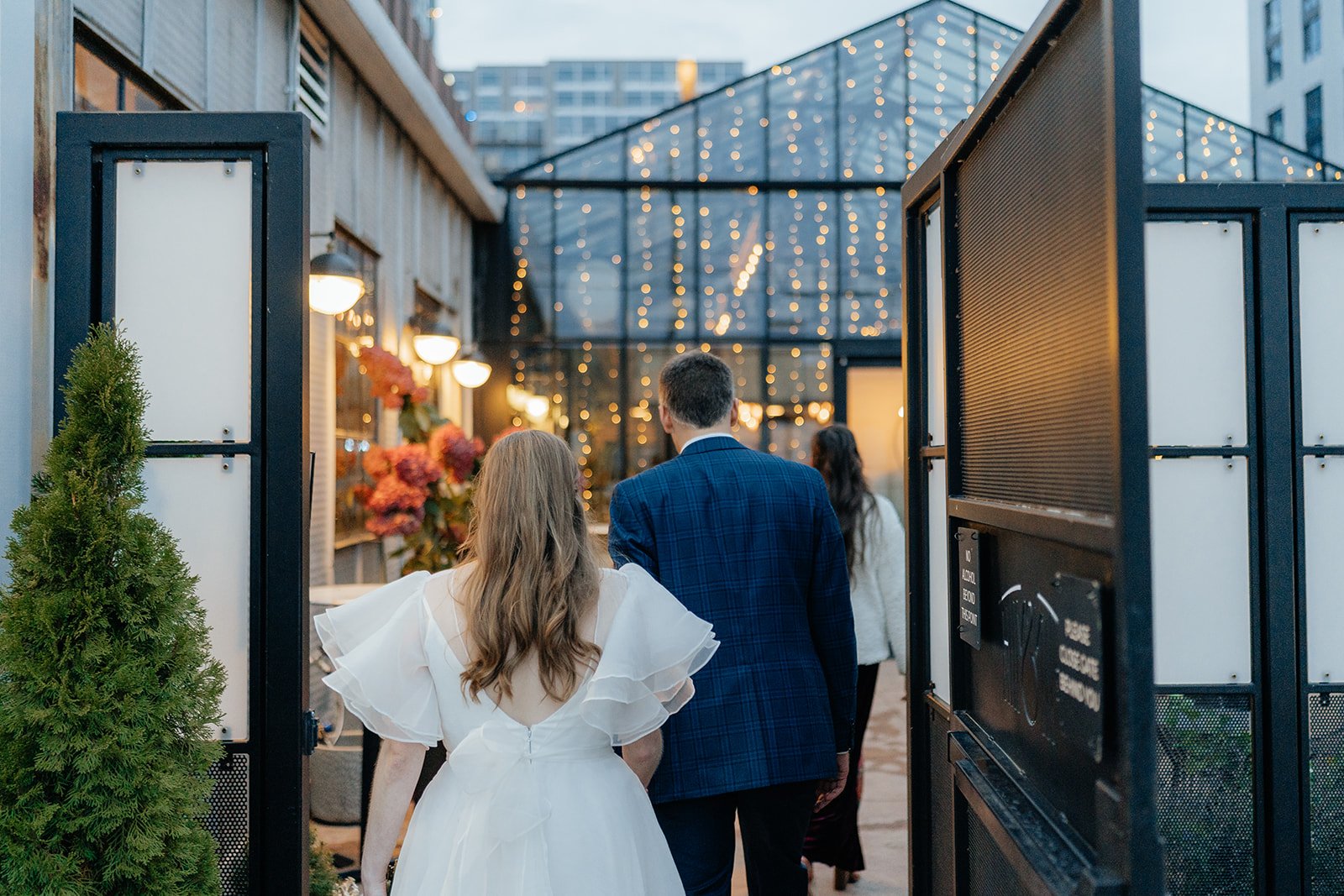 Bride and groom walking through the entrance of a modern venue with string lights and glass architecture, capturing the atmosphere of weddings Madison WI and stylish places to get married in Madison WI.