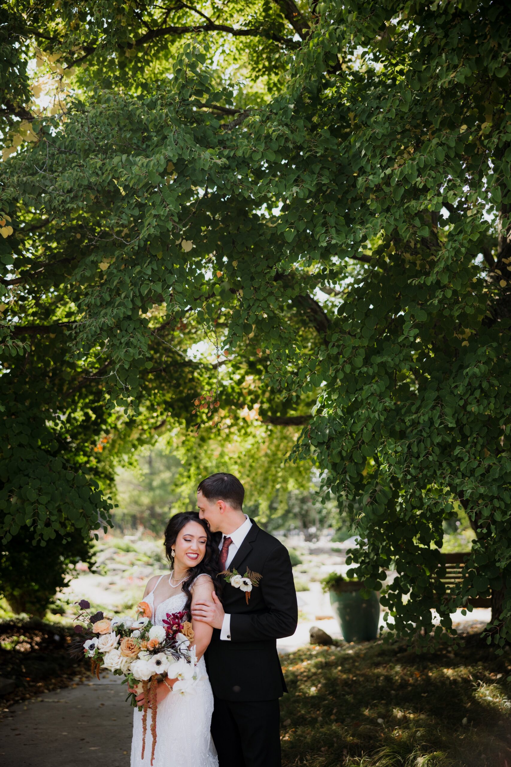 Bride and groom sharing a quiet moment beneath a leafy tree while holding a bouquet, capturing the romance of weddings Madison WI and scenic outdoor places to get married in Madison WI.