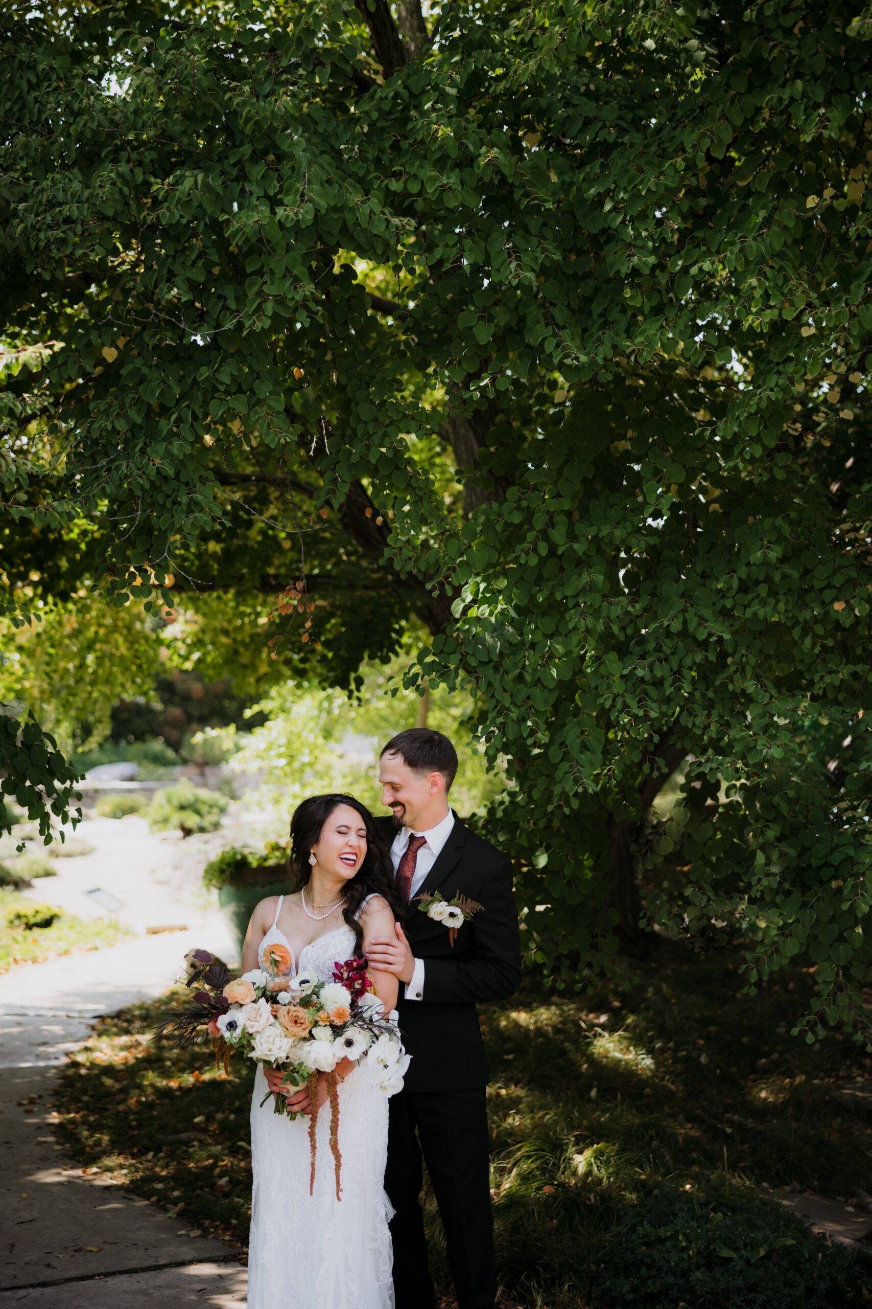 Bride and groom sharing a laugh beneath a leafy tree while holding a bouquet, capturing the romance of weddings Madison WI and scenic outdoor places to get married in Madison WI.